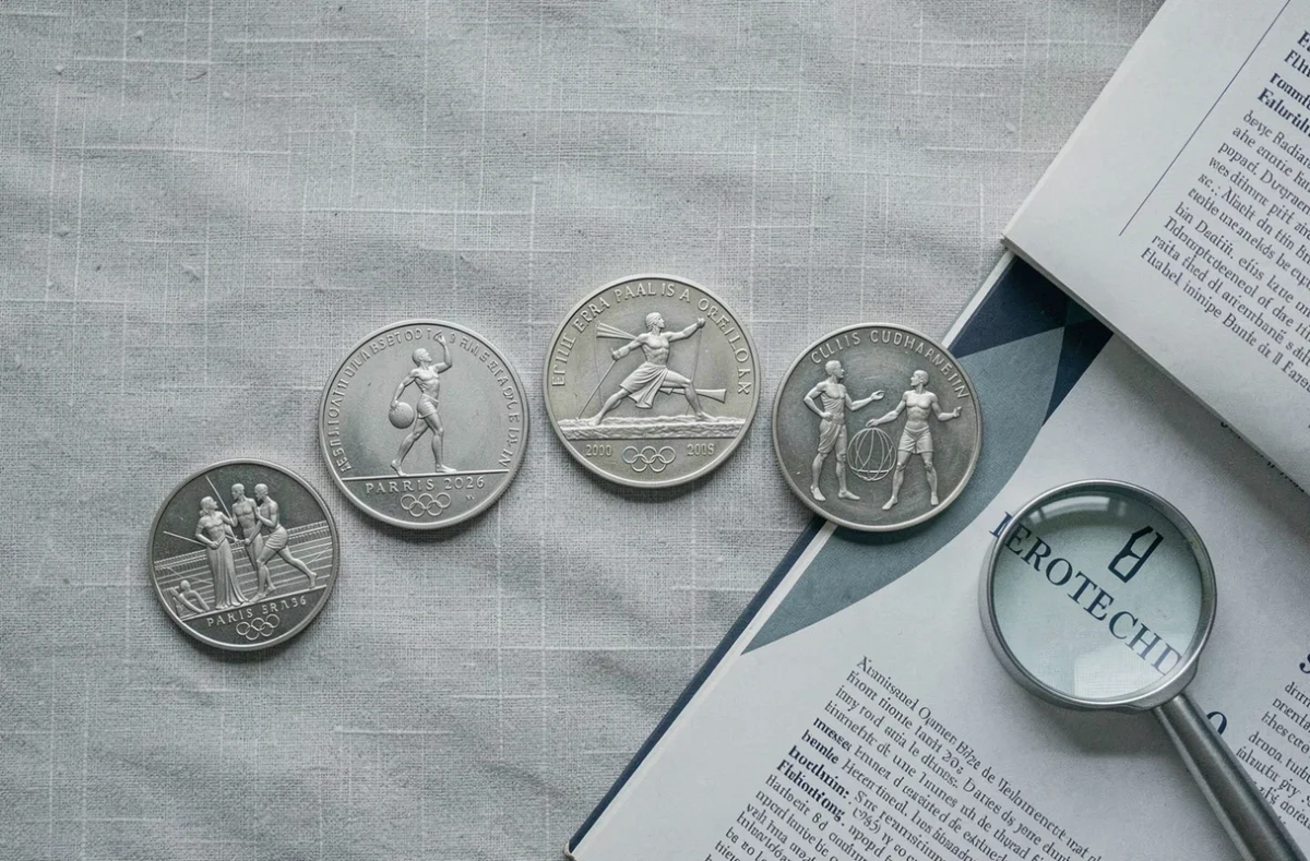Collection de pièces commémoratives euro Jeux Olympiques 2026 disposées en arc sur tissu lin avec loupe de collectionneur